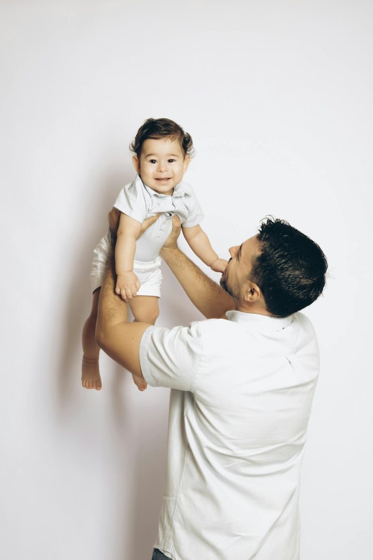 A father lovingly lifts his smiling baby boy against a white background, showcasing a joyful family moment.